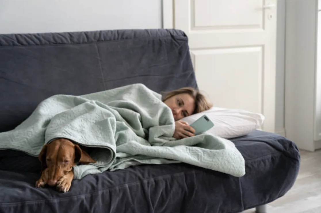 Woman resting under a blanket on the couch with her dog, checking her phone